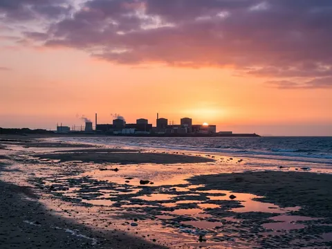 Une centrale nucléaire en bord de mer, vue depuis la plage à marée basse, sous un ciel orange et rose.