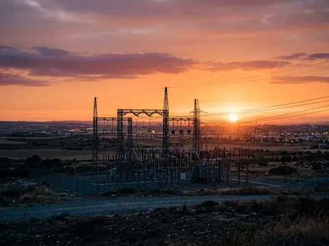 Un poste électrique très haute tension en silhouette au coucher du soleil, les lumières d'une ville qui s'allument au loin.
