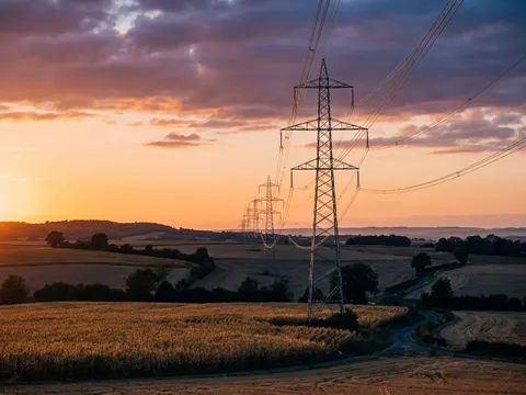 Des pylônes à très haute tension en enfilade au-dessus de champs, sous un ciel orange et mauve de fin de journée.